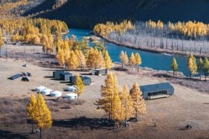 Aerial view of Khanagai Lodge on the Shishged River in northern Mongolia, showing riverside cabins, Mongolian gers, and the main lodge surrounded by autumn larch forest